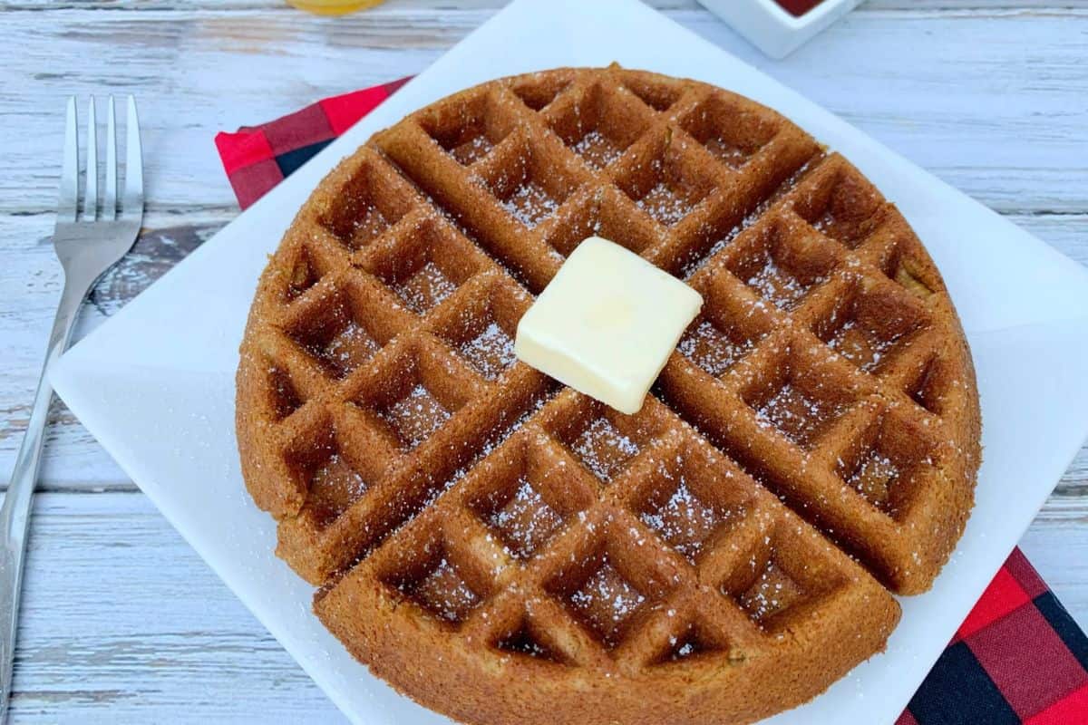 A round waffle topped with a pat of butter and dusted with powdered sugar sits on a white plate next to a fork and plaid napkin—perfect for a cozy Christmas morning breakfast.