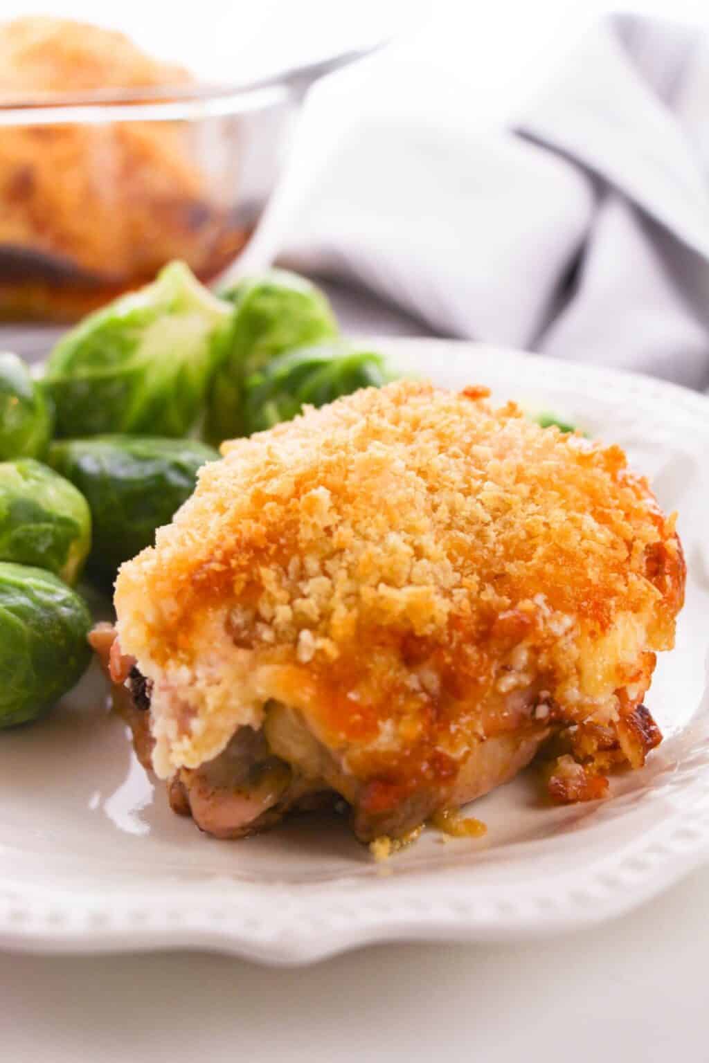 A Hellman’s Crusted Parmesan Chicken thigh with a golden, crispy crust is served on a white plate alongside steamed Brussels sprouts.