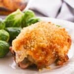 A Hellman’s Crusted Parmesan Chicken thigh with a golden, crispy crust is served on a white plate alongside steamed Brussels sprouts.