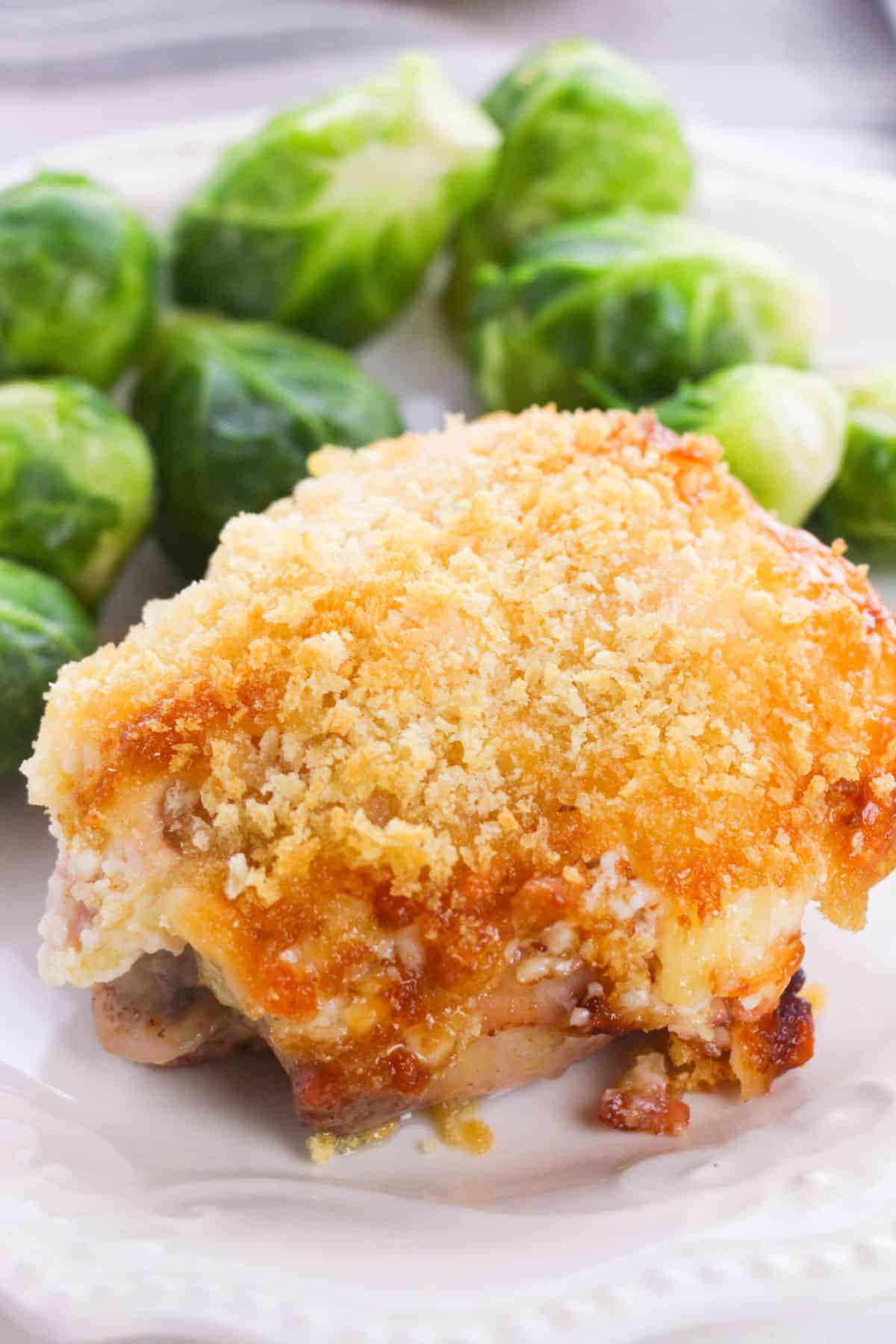 A Hellman's Crusted Parmesan Chicken thigh, breaded and baked to perfection, is served on a white plate alongside several steamed Brussels sprouts.