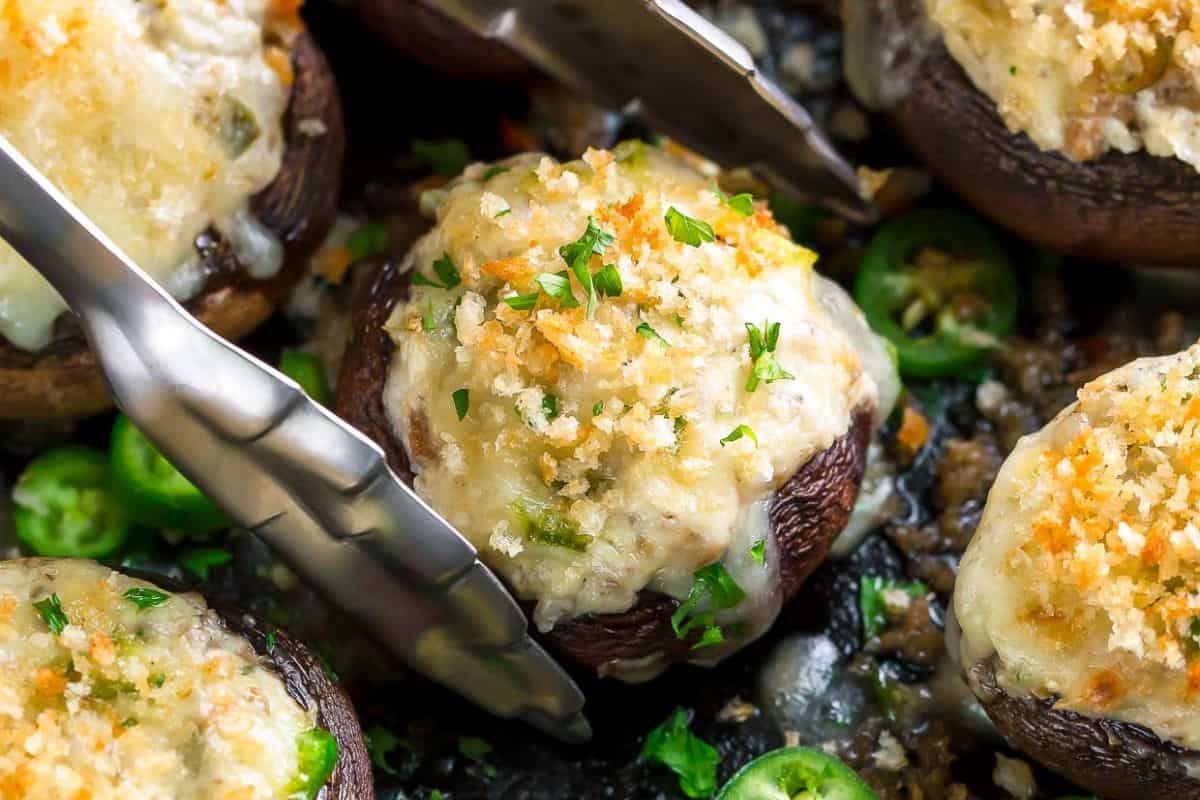 Stuffed mushrooms topped with melted cheese and breadcrumbs are held by metal tongs, with sliced green peppers and herbs scattered around—perfect for anyone searching for delicious stuffed mushroom recipes.
