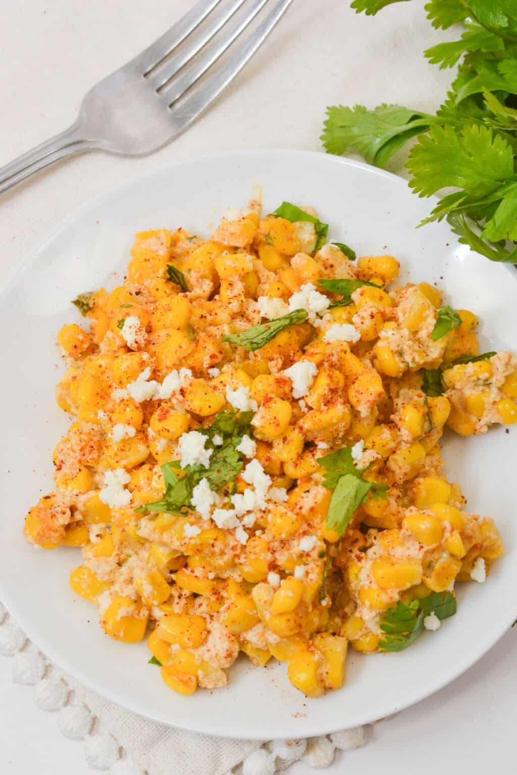 A plate of seasoned Mexican Street Corn Casserole topped with crumbled cheese and fresh cilantro, with a fork and cilantro leaves nearby on a light tablecloth.