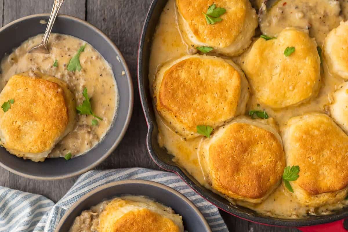 A cast iron skillet with golden biscuits in creamy gravy, next to a bowl of biscuits and gravy, garnished with parsley—perfect for a cozy Christmas recipe or festive holiday breakfast.