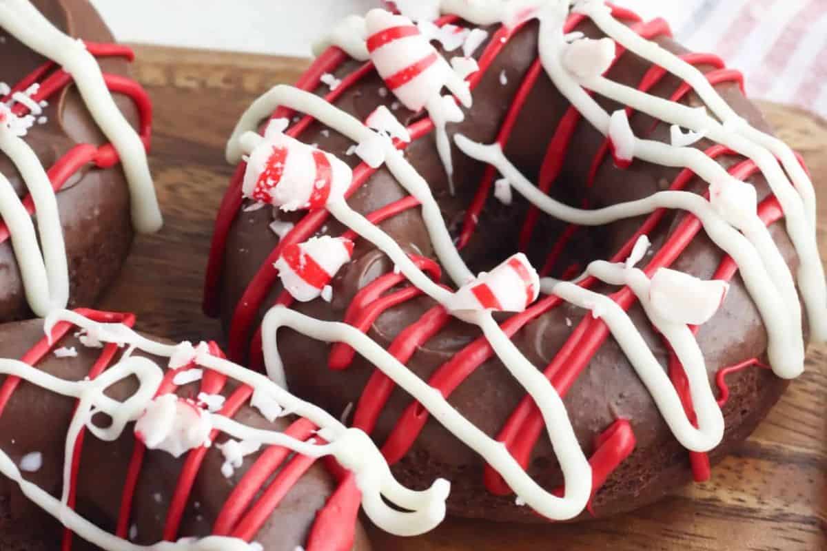Chocolate donuts topped with red and white icing drizzles and sprinkled with crushed peppermint candy—perfect for a cozy Christmas morning breakfast on a wooden surface.