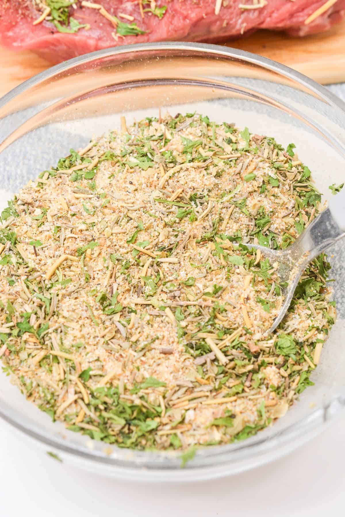 A clear glass bowl filled with a mixed dry herb and spice pot roast seasoning recipe blend, with a spoon partially visible on the right side.