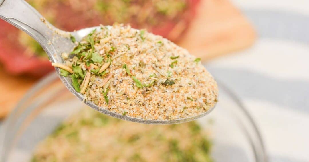 A close-up of a spoon holding a mixed dry Pot Roast Seasoning recipe with herbs and spices, above a bowl containing more of the mixture.