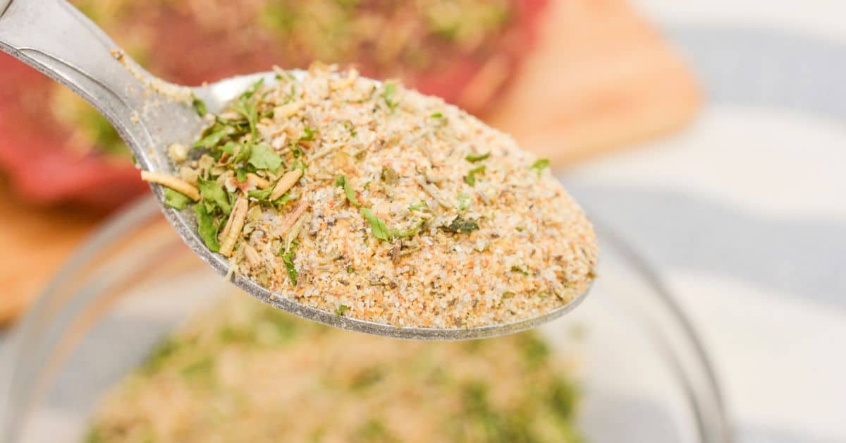 A close-up of a spoon holding a mixed dry Pot Roast Seasoning recipe with herbs and spices, above a bowl containing more of the mixture.