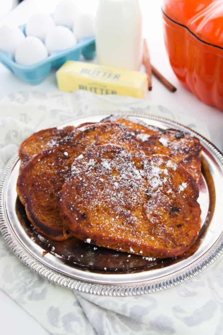 Slices of Pumpkin Pie Spice French Toast dusted with powdered sugar on a silver platter, with eggs, a stick of butter, a bottle of milk, and an orange pot in the background.