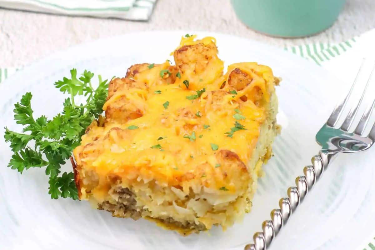 A slice of tater tot casserole topped with melted cheese and herbs, served on a white plate with parsley and a fork—perfect for cozy Christmas breakfast gatherings.