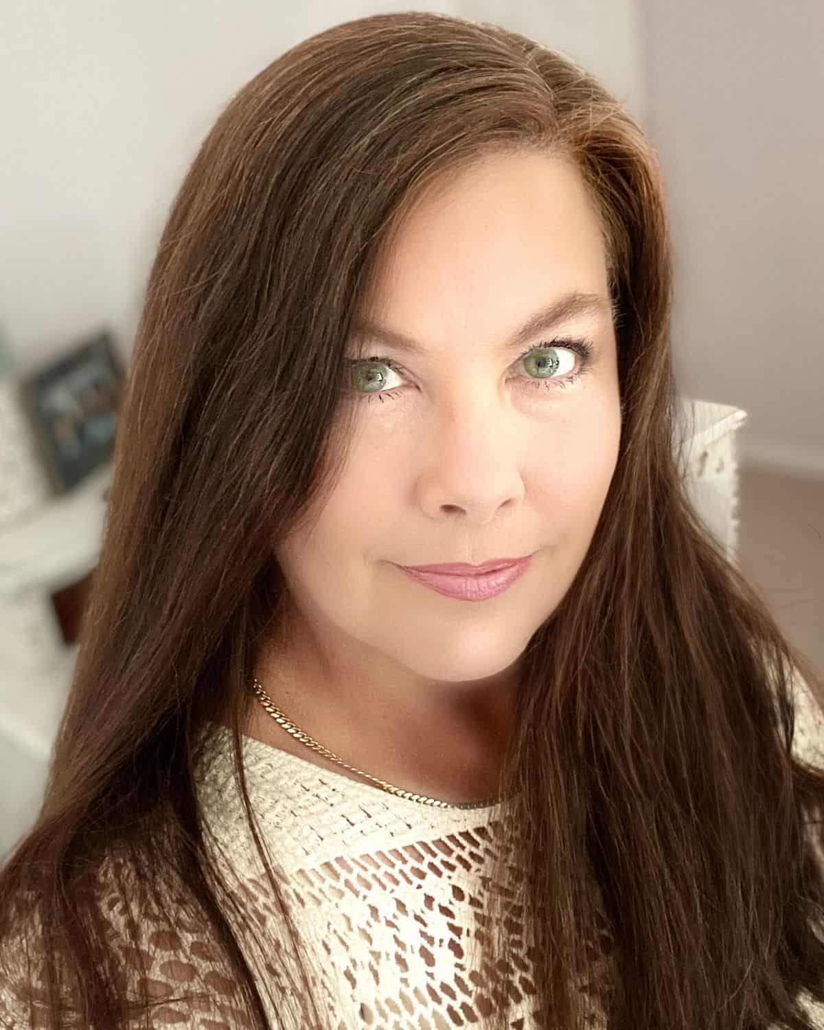 A woman with long brown hair and green eyes looks at the camera, wearing a white textured top and a gold necklace, indoors with a blurred background—ready to share her favorite Instant Pot recipes.
