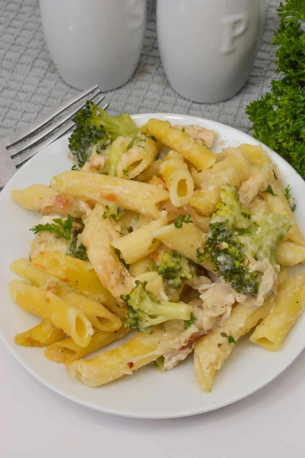 A plate of Baked Chicken and Broccoli Alfredo penne pasta with melted cheese, garnished with parsley, on a white plate with a fork nearby.