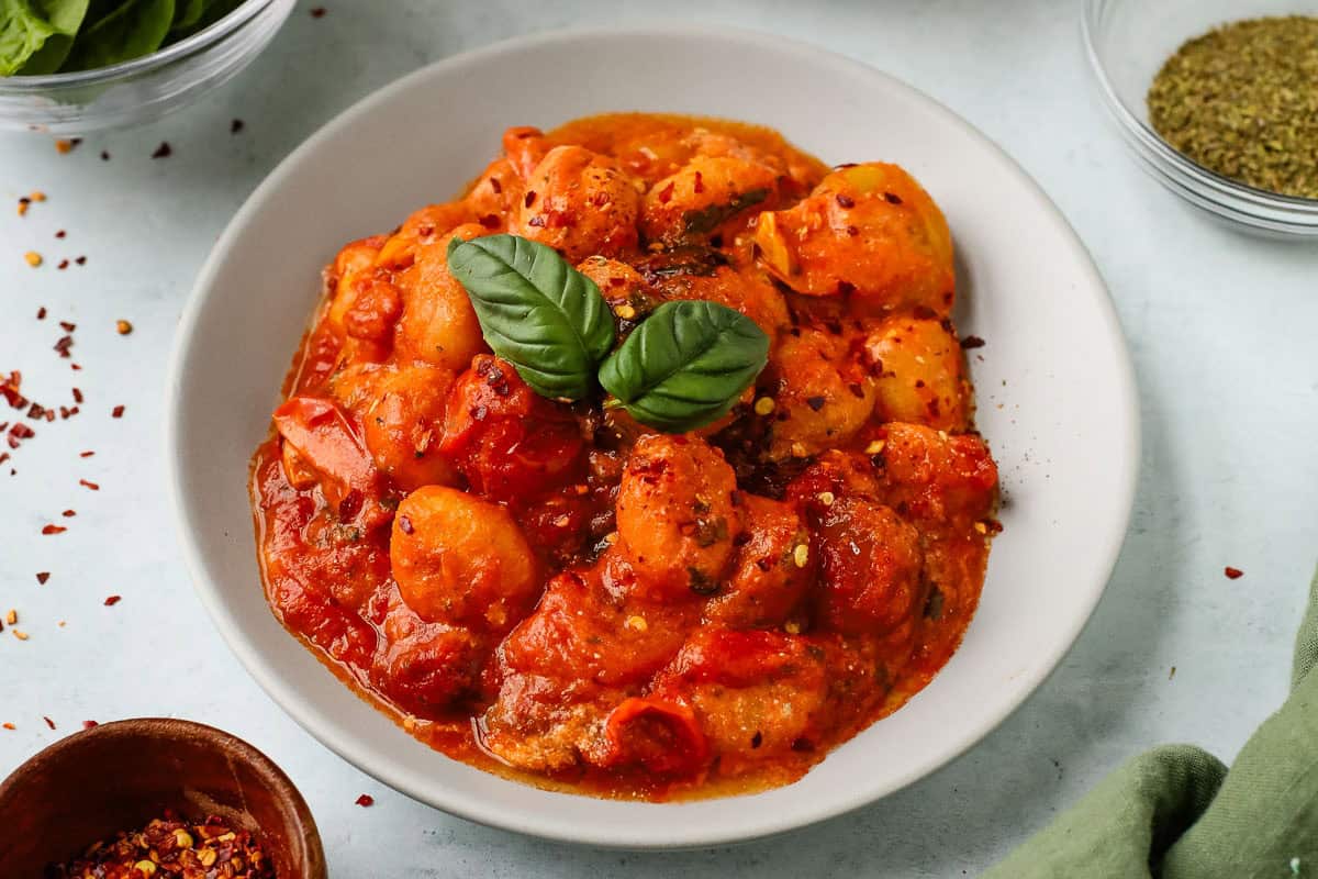 A bowl of gnocchi in tomato sauce is garnished with basil leaves and red pepper flakes sits on a white plate surrounded by seasonings and herbs.