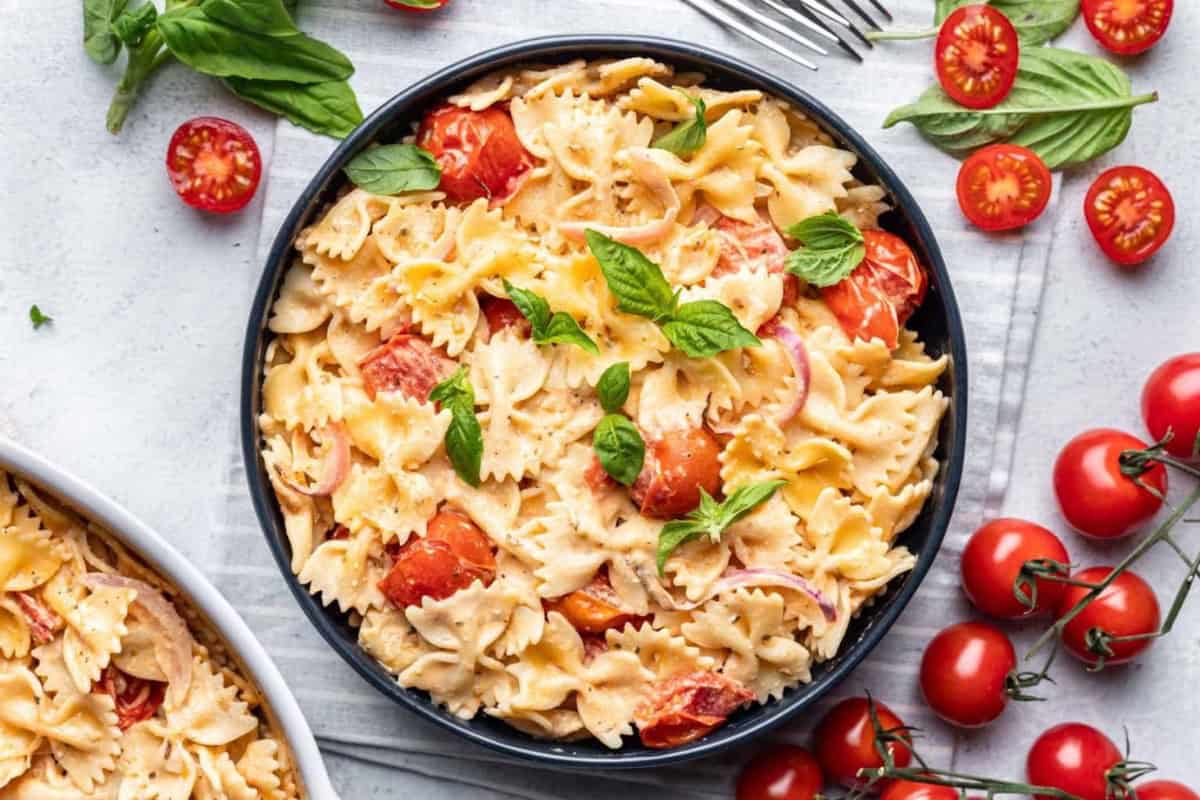 A bowl of Boursin cheese farfalle pasta mixed with cherry tomatoes, red onions, and basil leaves surrounded by fresh tomatoes and basil on a white surface.