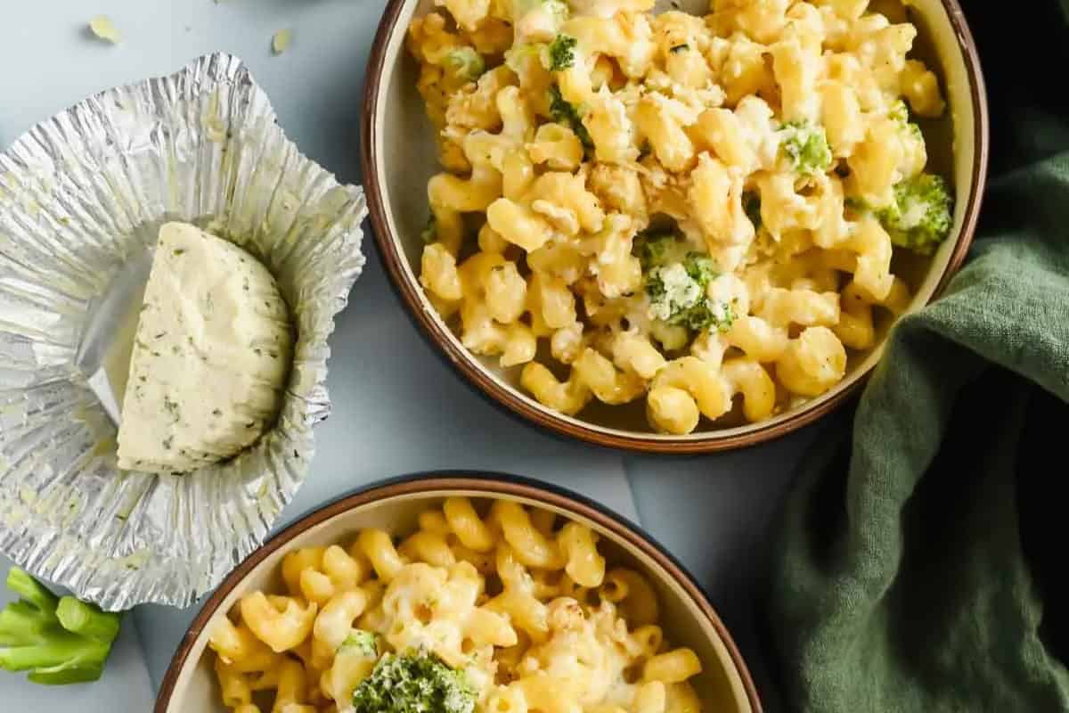 Two bowls of Boursin macaroni and cheese with broccoli sit beside a foil-wrapped portion of Boursin cheese and a green cloth napkin.