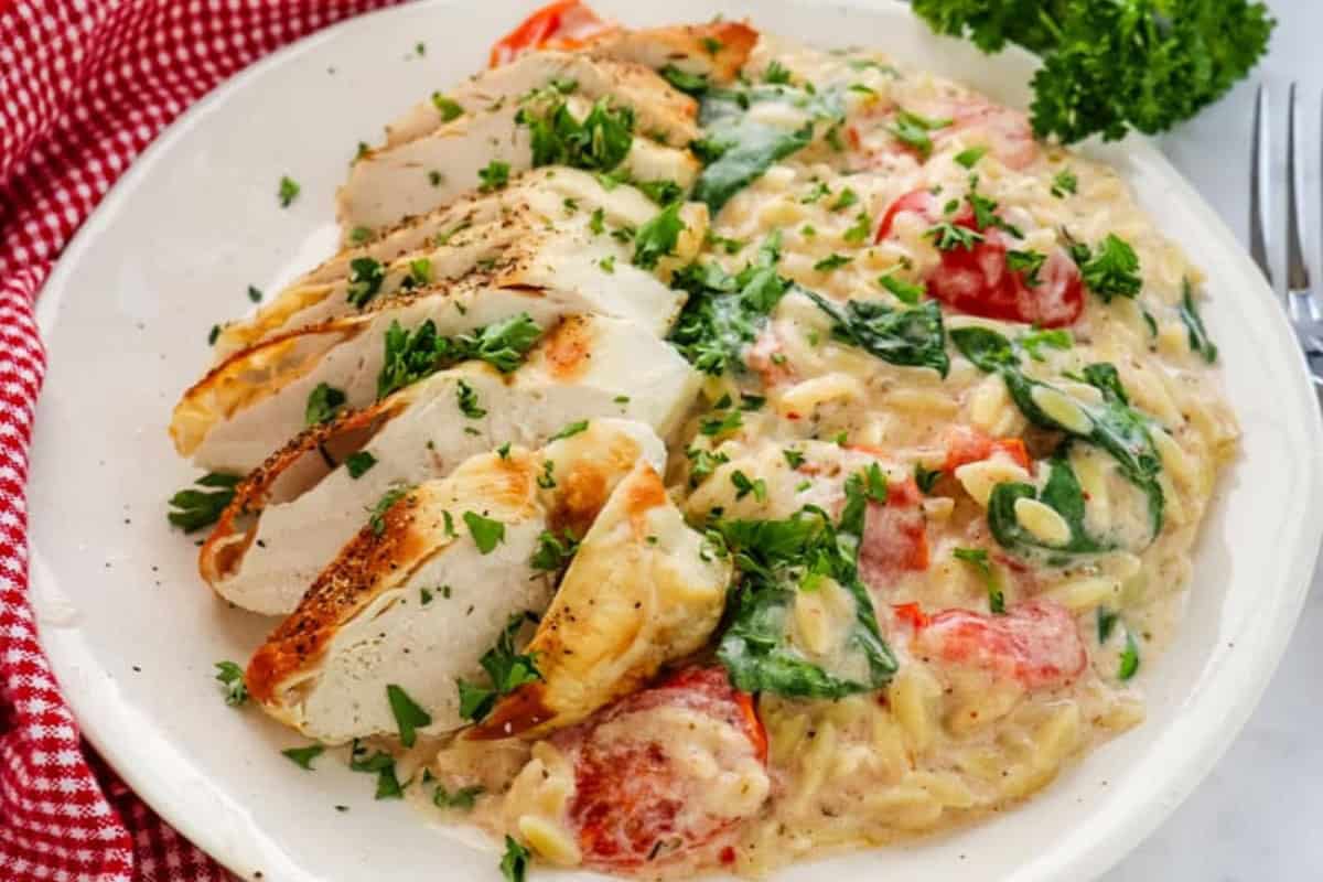 Sliced grilled chicken breast served with creamy Boursin cheese orzo pasta, spinach, and tomatoes on a white plate, garnished with herbs.