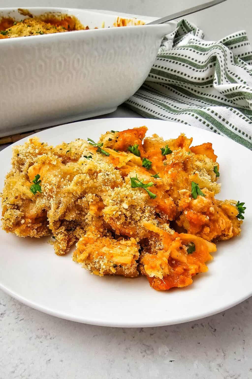 A serving of Chicken Parmesan Casserole with a golden breadcrumb topping and parsley on a white plate, with a casserole dish and striped cloth in the background.