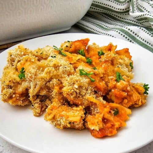 A serving of Chicken Parmesan Casserole with a golden breadcrumb topping and parsley on a white plate, with a casserole dish and striped cloth in the background.
