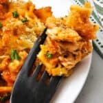 A close-up of a black fork holding a bite of baked pasta with sauce and herbs—above a white plate of the same dish. A striped cloth is visible in the background.