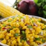 A bowl of corn salad with chopped red onion and green herbs, inspired by a Chipotle Corn Salsa Recipe, with an ear of corn, a red onion, and cilantro in the background.