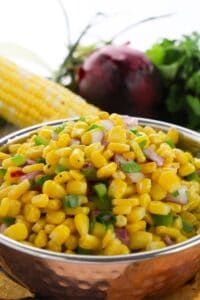 A bowl of corn salad with chopped red onion and green herbs, inspired by a Chipotle Corn Salsa Recipe, with an ear of corn, a red onion, and cilantro in the background.