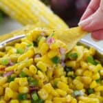 A hand holds a tortilla chip scooping Chipotle Corn Salsa Recipe with diced red onion, jalapeño, and cilantro from a metal bowl. An ear of corn and red onion are in the background.