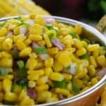 A close-up of a bowl filled with corn salad mixed with chopped red onions, cilantro, and green peppers.. Corn on the cob and fresh vegetables are in the background.