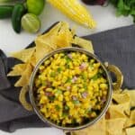 A bowl of the side with diced red onion and green peppers, surrounded by tortilla chips, with corn, jalapeño, lime, red onion, and cilantro in the background.