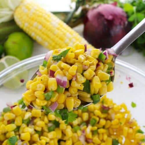 A spoonful of Chipotle Corn Salsa Recipe with red onion and herbs is held above a bowl, with fresh corn, lime, and vegetables in the background.