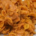 A close-up of Cornflake Cookies showcases clusters of cornflakes coated in a glossy, caramel-colored peanut butter mixture on a white plate.