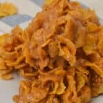 A close-up of a stack of caramel-coated Cornflake Cookies clusters on a white plate, with a striped fabric in the background.