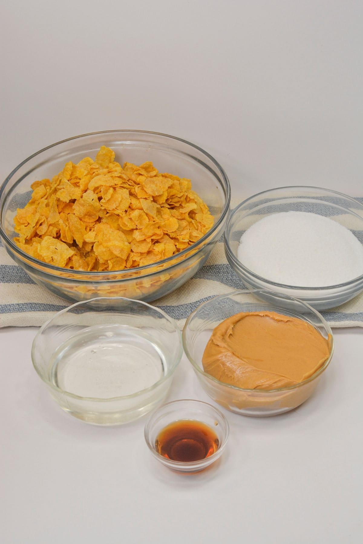 Five glass bowls containing the ingredients cereal, granulated sugar, peanut butter, light corn syrup, and vanilla extract—are arranged on a striped cloth against a plain background.