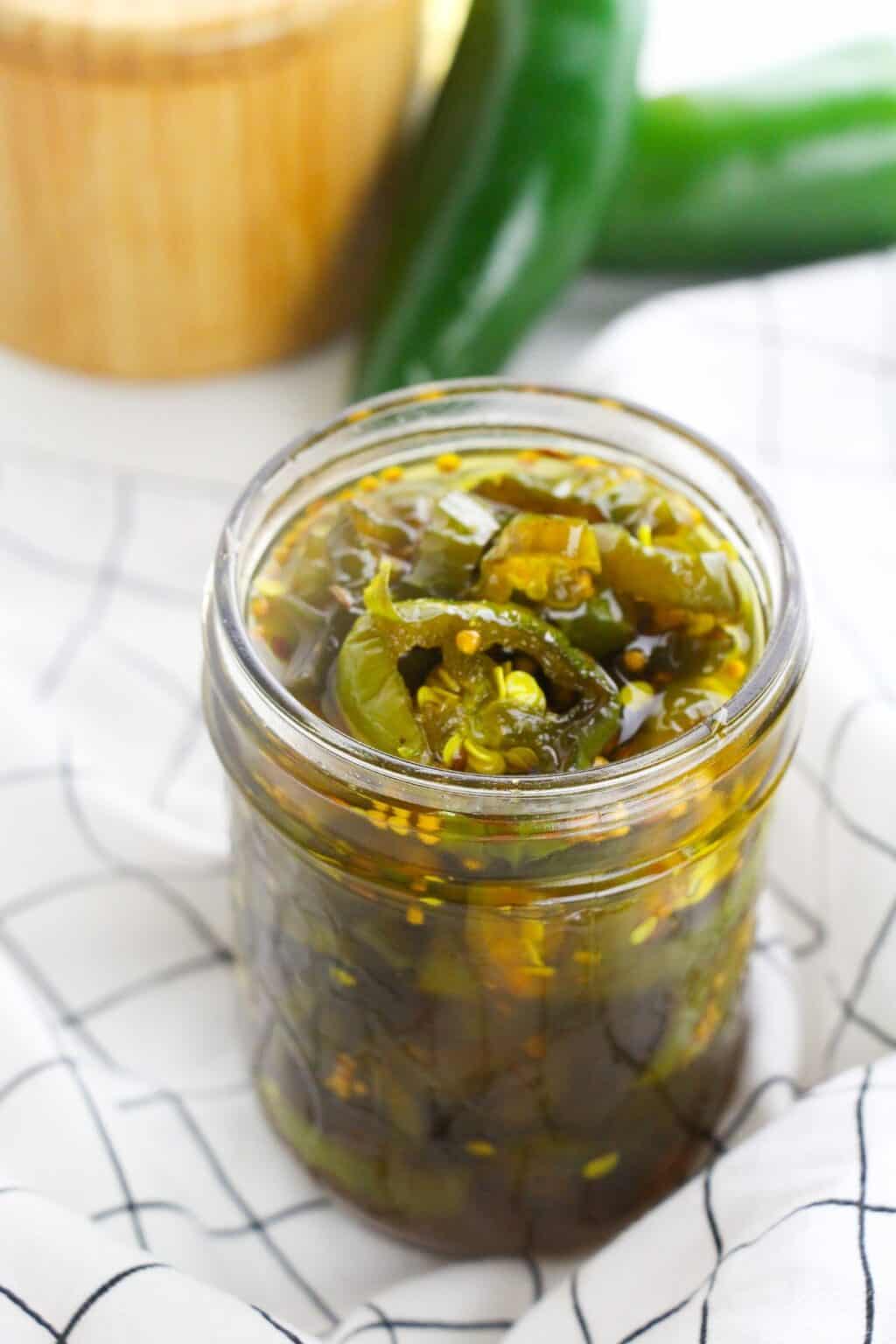 A glass jar filled with sliced Cowboy Candy pickled jalapeños in liquid, placed on a white cloth with a black grid pattern.