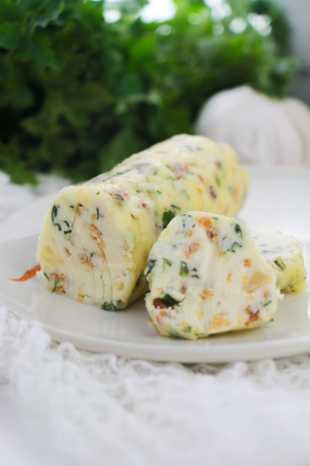 A log of Garlic Herb Steak Butter with herbs and vegetables is sliced on a white plate, with greenery in the blurred background.