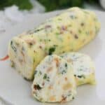 A log of Garlic Herb Steak Butter with herbs and chopped ingredients sits on a white plate, with two slices cut from it.