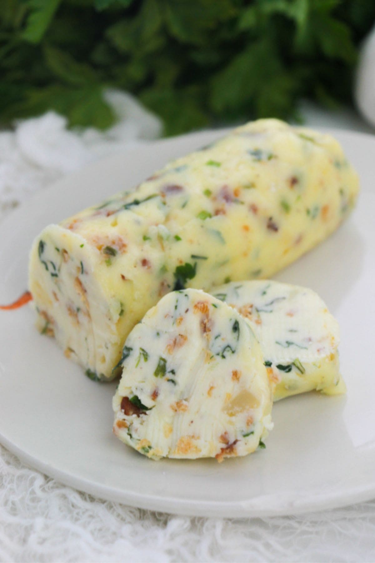 A log of Garlic Herb Steak Butter with herbs and chopped ingredients sits on a white plate, with two slices cut from it.