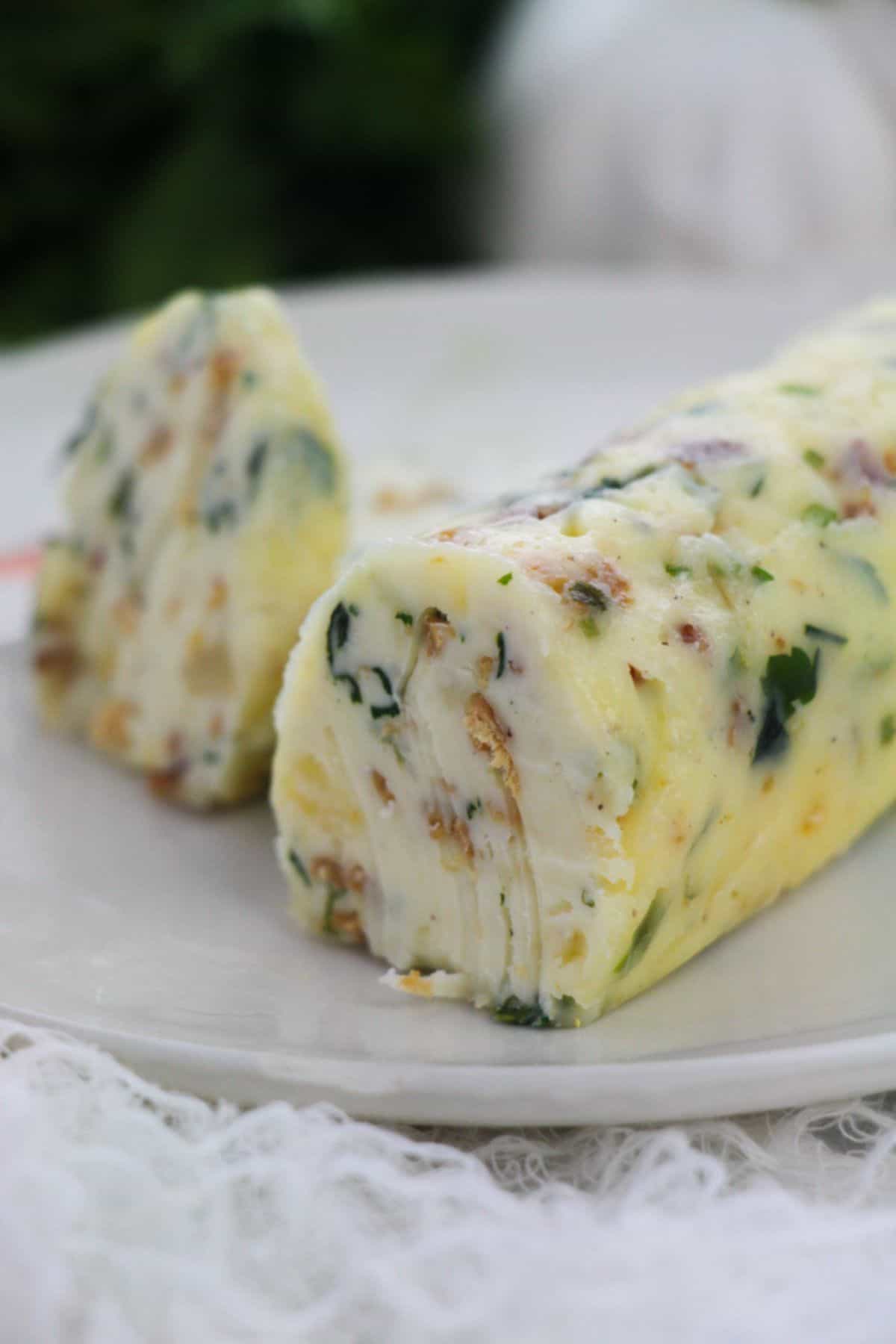 A log of garlic herb steak butter, flecked with herbs and bits of nuts or bacon, sits on a white plate, with one slice cut from the end.