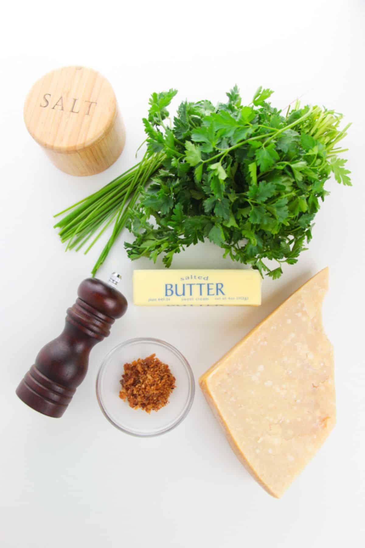 A bunch of parsley and chives, a stick of salted butter, a chunk of Parmesan, a salt container, a pepper grinder, and a small bowl of brown seasoning—perfect ingredients are arranged on a white background.