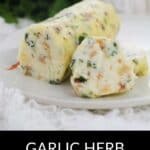 A log of Garlic Herb Steak Butter with visible herbs and spices, partially sliced on a white plate, surrounded by greenery and a garlic bulb. Text reads: Garlic Herb Steak Butter.