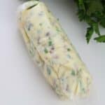 A log of Garlic Herb Steak Butter with herbs and spices, partially wrapped in parchment paper, placed on a white surface next to fresh parsley.