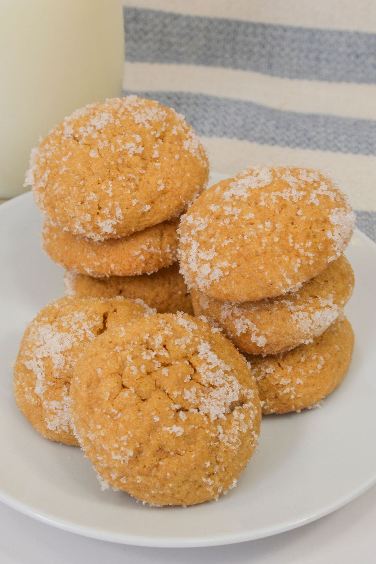 A white plate with stacks of round, golden Ginger Molasses Cookies coated in granulated sugar, set in front of a blue and white striped cloth.