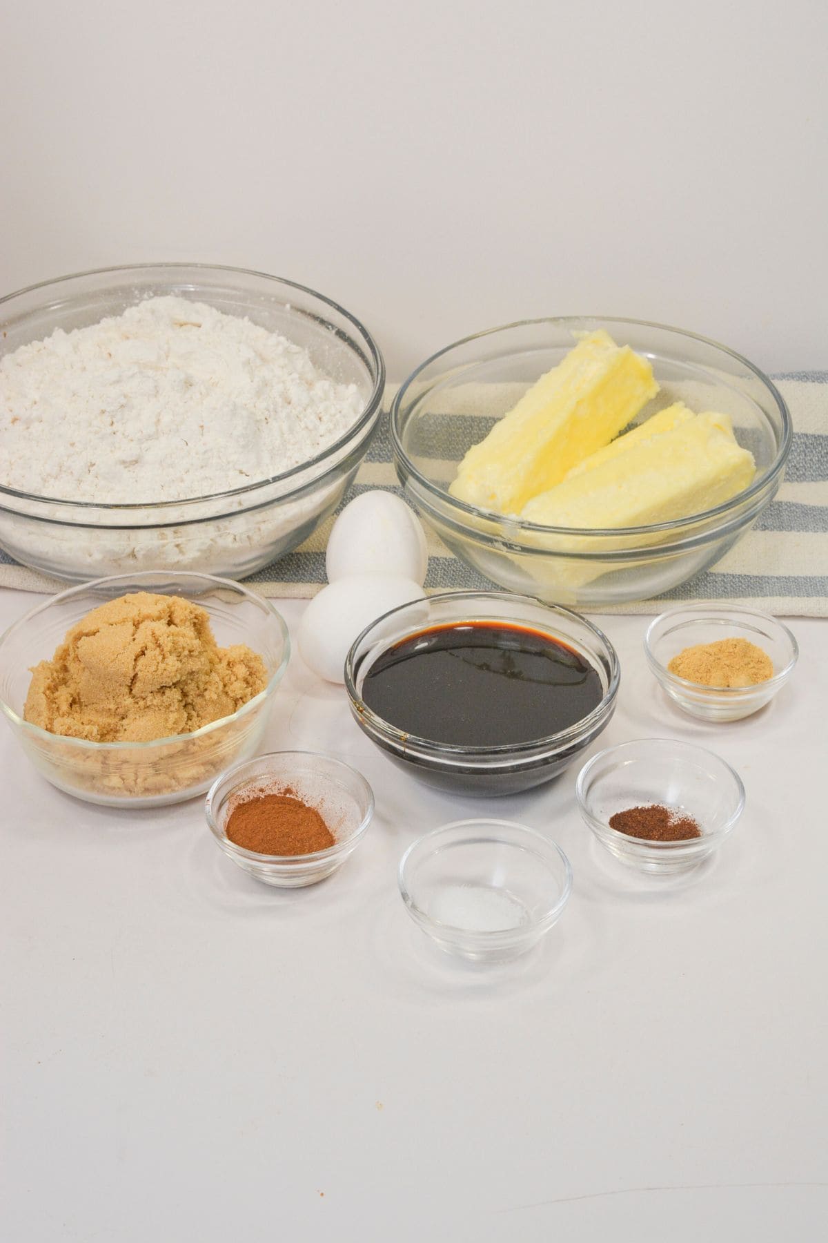 A variety of baking ingredients for Ginger Molasses Cookies are arranged in clear bowls, including flour, brown sugar, butter, eggs, molasses, and several spices on a white surface.