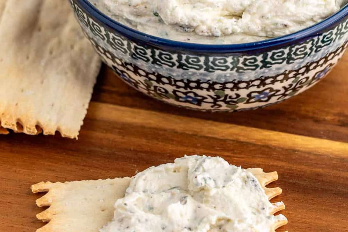 A blue and white patterned bowl of creamy white homemade Boursin dip with garlic and herbs sits on a wooden surface next to a rectangular cracker spread with the dip. Additional plain crackers sit to the left side of the bowl.