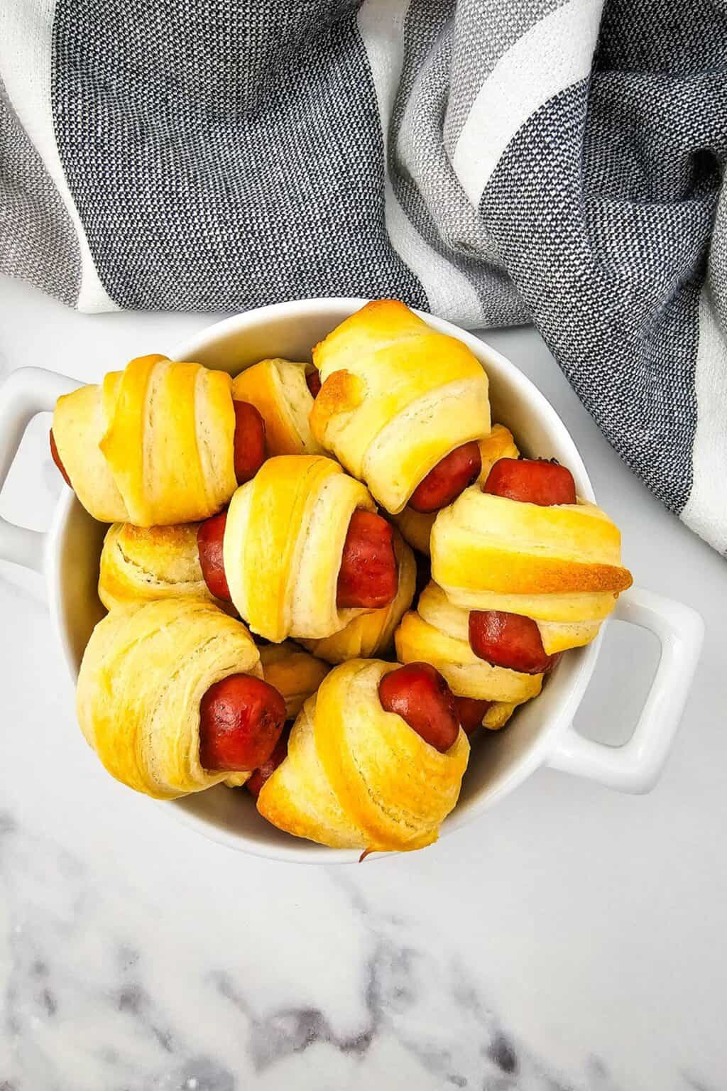 A white dish filled with several Mini Pigs in a Blanket, small sausages wrapped in golden-brown pastry, sits on a marble surface next to a striped cloth.