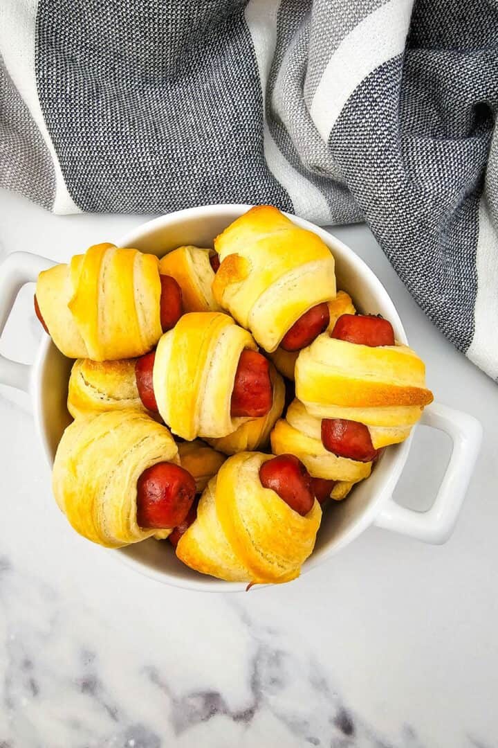 A white dish filled with several Mini Pigs in a Blanket, small sausages wrapped in golden-brown pastry, sits on a marble surface next to a striped cloth.
