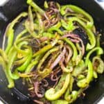 Sautéed onions and peppers—green bell peppers and red onions—sizzle in a cast iron skillet, slightly charred, with a blue patterned cloth in the background.
