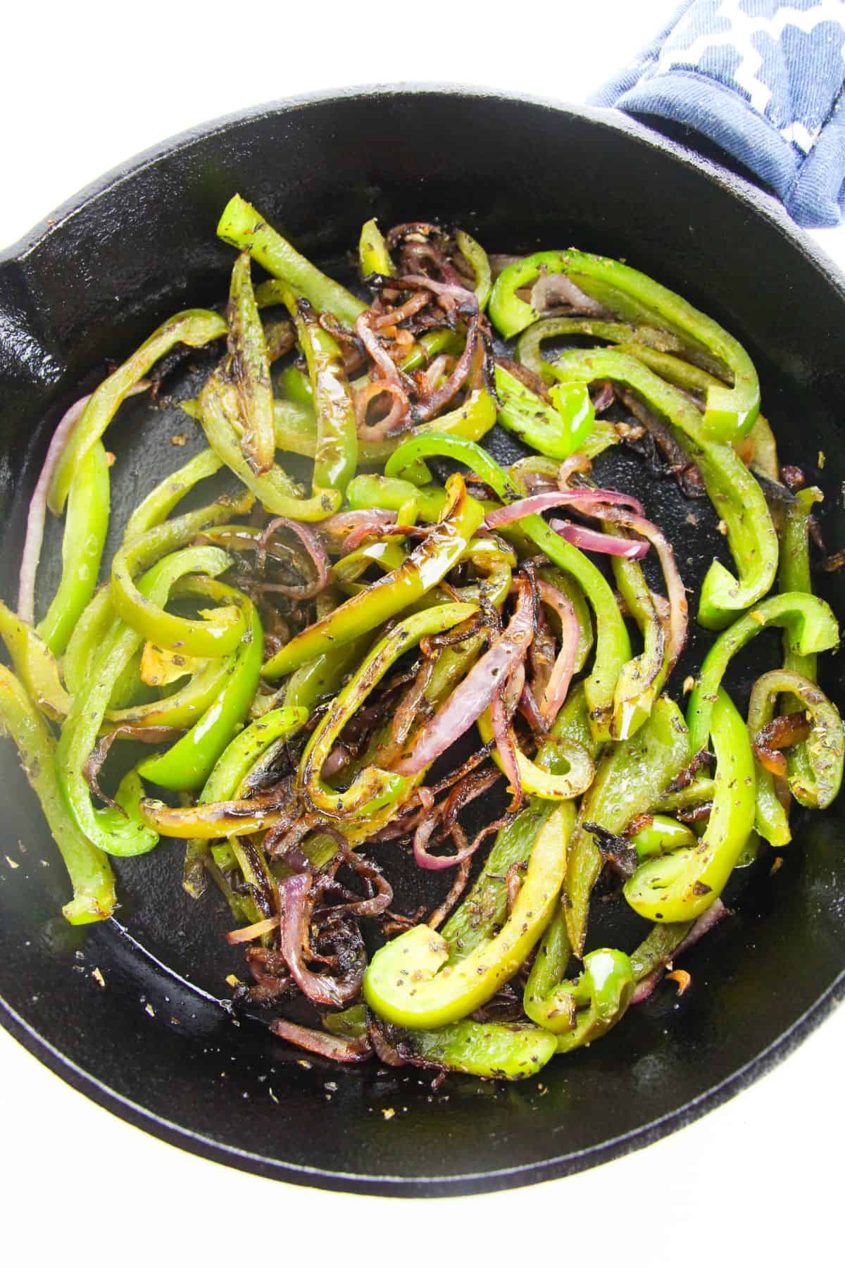 Sautéed onions and peppers—green bell peppers and red onions—sizzle in a cast iron skillet, slightly charred, with a blue patterned cloth in the background.
