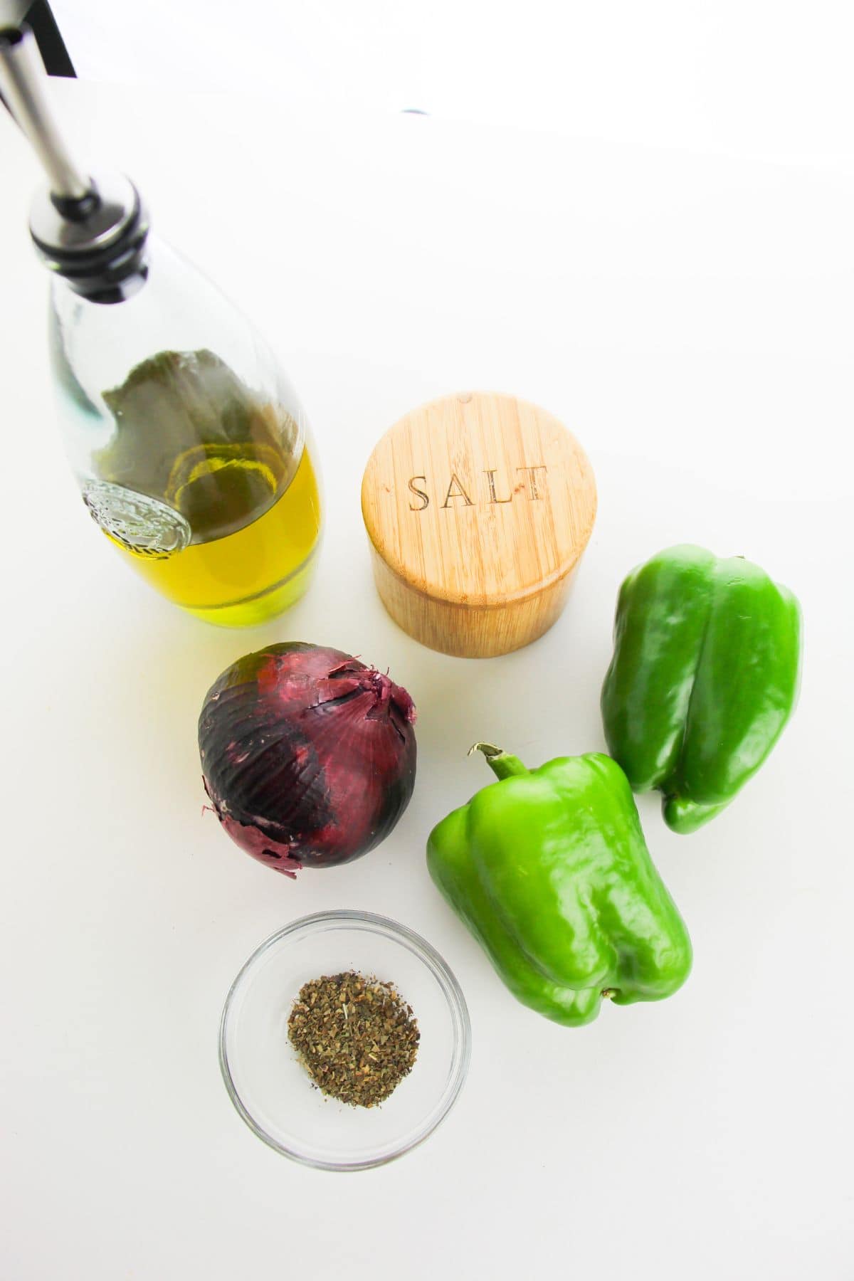 A bottle of olive oil, a wooden salt container, a red onion, two green bell peppers, and a small bowl of dried herbs are arranged on a white surface—perfect ingredients for delicious Sautéed Onions and Peppers.