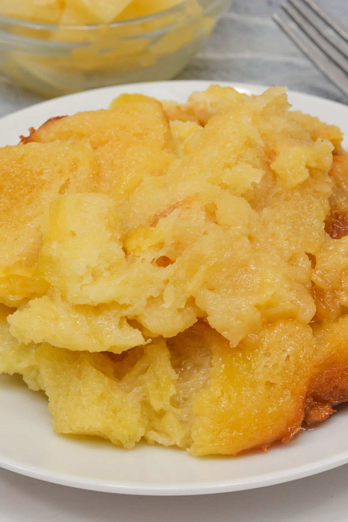 A serving of Pineapple Casserole on a white plate, with a bowl of pineapple chunks and a fork in the background.