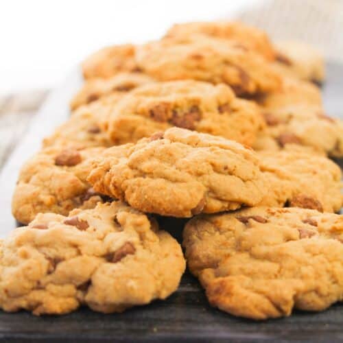 A row of soft chocolate chip cookies is arranged on a dark serving board with a printed cloth underneath.
