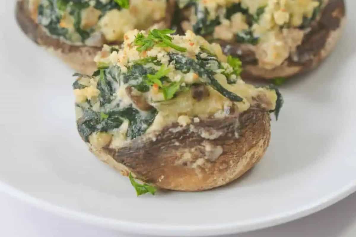 A stuffed mushroom cap filled with a savory mixture of spinach, Boursin Cheese, breadcrumbs and shallots, served on a white plate. Two other stuffed mushrooms can be seen blurred in the background behind the one in the front.